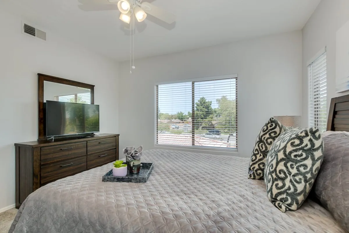 A cozy bedroom with a comfortable bed adorned with decorative pillows, a dresser with a television, and large windows with blinds letting in soft natural light. The space feels both restful and functional, inviting relaxation and entertainment.