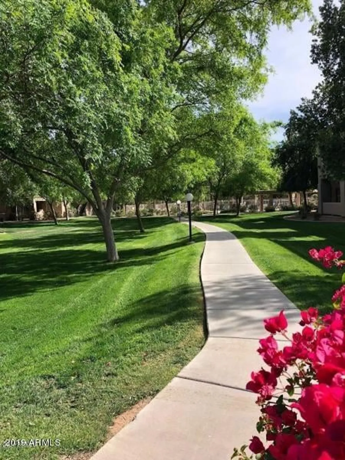 Beautifully landscaped community grounds featuring a winding pathway, flowering plants, mature trees, and a classic lamp post. The scene is peaceful and encourages leisurely strolls or moments of relaxation amidst nature.