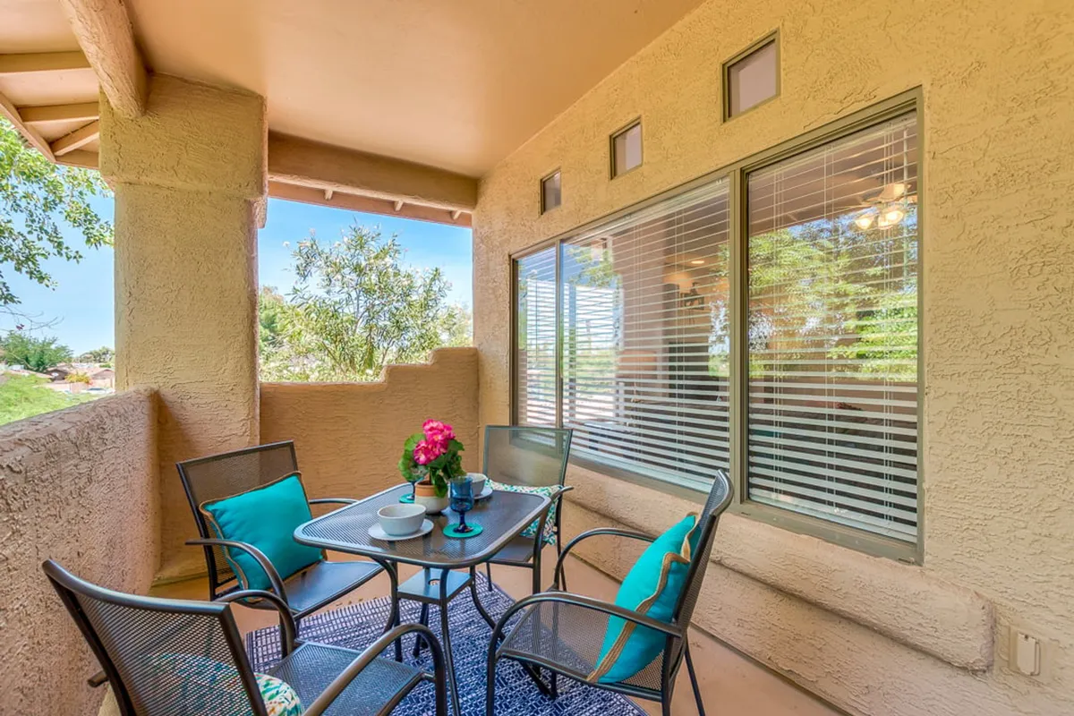 A cozy balcony or patio scene featuring a table with fresh flowers, colorful cushions, and a large window backdrop. The comfortable setting is perfect for both morning relaxation and evening gatherings, reinforcing the concept of extending the living space outdoors.