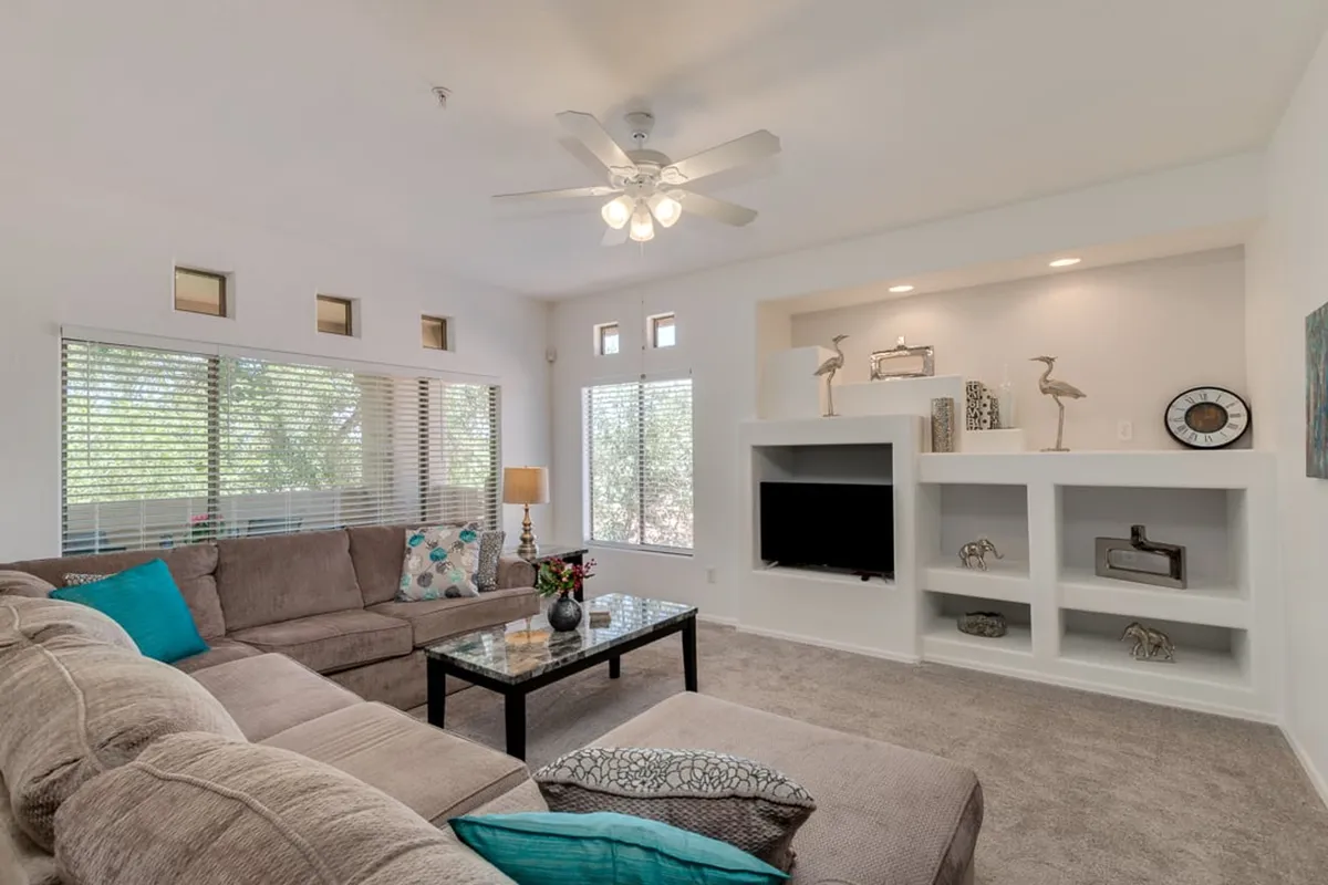 A bright, spacious living room with a comfortable sofa, coffee table, decorative shelves, and large windows letting in ample natural light. The area is styled for relaxation or productivity, with a versatile layout ideal for unwinding after work or catching up on emails.