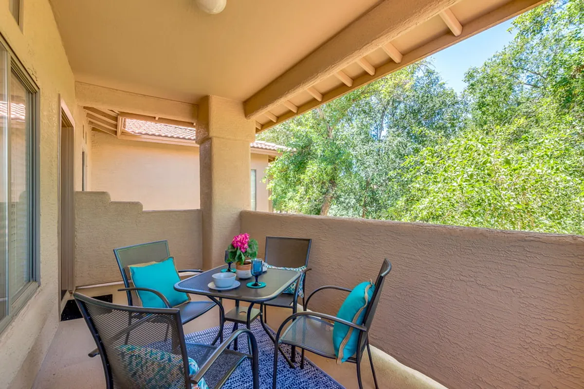 An inviting exterior shot of a balcony or patio area with a table and chairs surrounded by potted flowers and abundant lush greenery. The scene suggests an intimate setting for enjoying morning coffee or evening gatherings under the open sky.