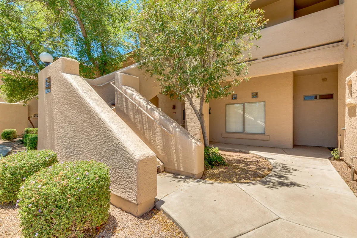 This exterior image shows a welcoming staircase leading to the condo entrance, bordered by a leafy tree and a well-maintained pathway. The scene features natural light, inviting greenery, and a sense of calm, hinting at the community atmosphere business travelers may enjoy.