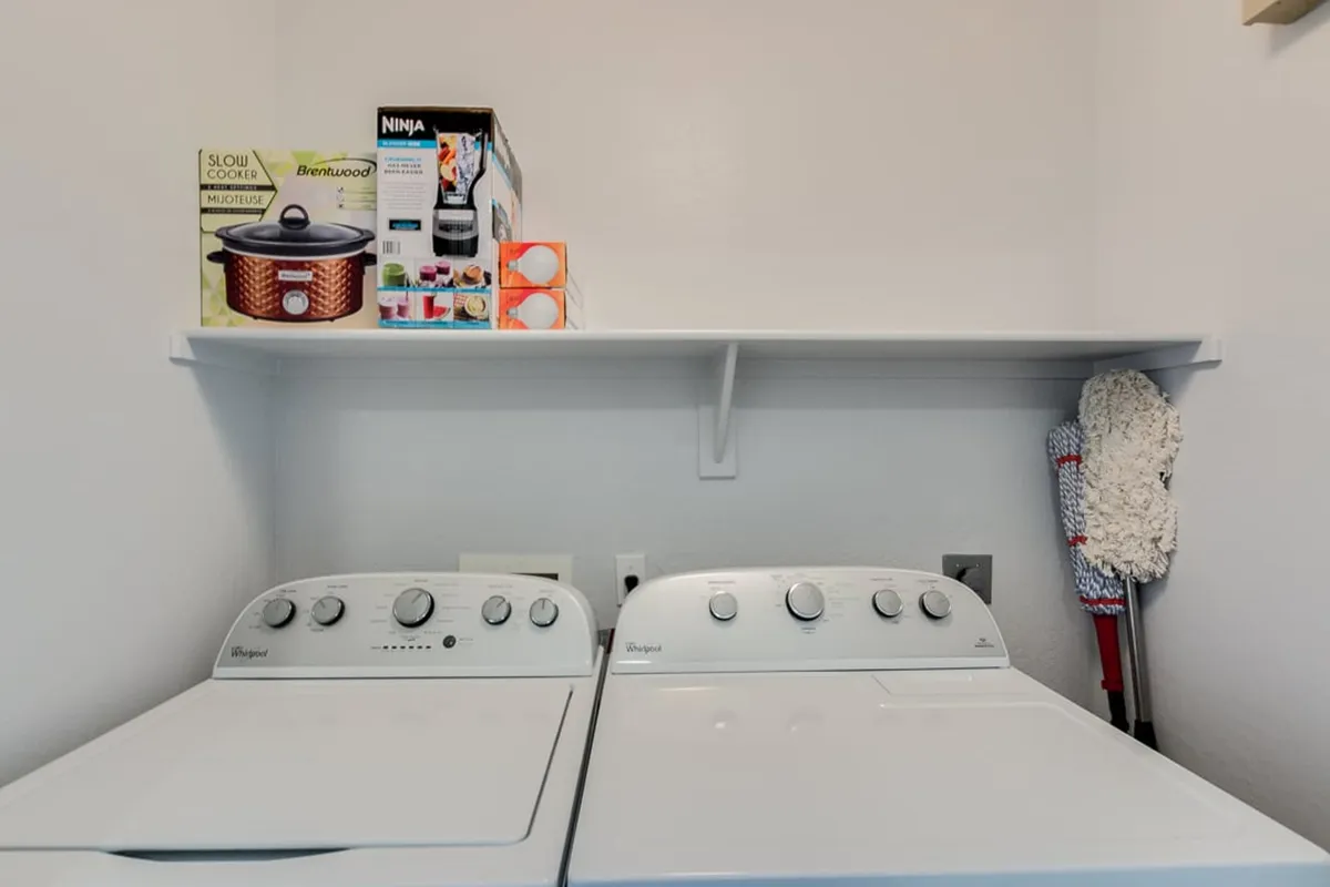 A practical in-unit laundry area with a modern washing machine and dryer beneath open shelving holding kitchen appliances. The space is tidy and efficient—ideal for keeping up with daily chores conveniently at home.