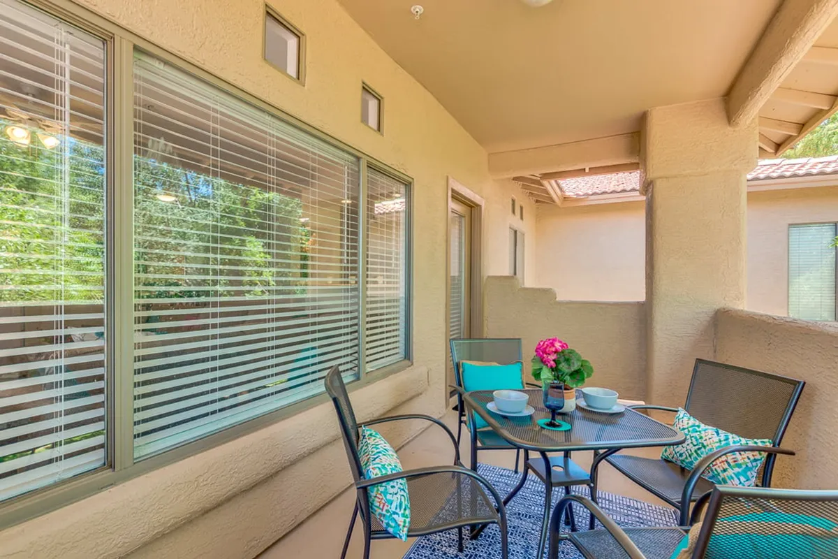 This is an exterior image of an inviting outdoor seating area with a table adorned by a flower arrangement. Large windows connect the outdoor space visually to the interior, offering a glimpse of a welcoming home environment. The setup is ideal for enjoying coffee, relaxing with friends, or hosting a small brunch while immersed in the neighborhood atmosphere.