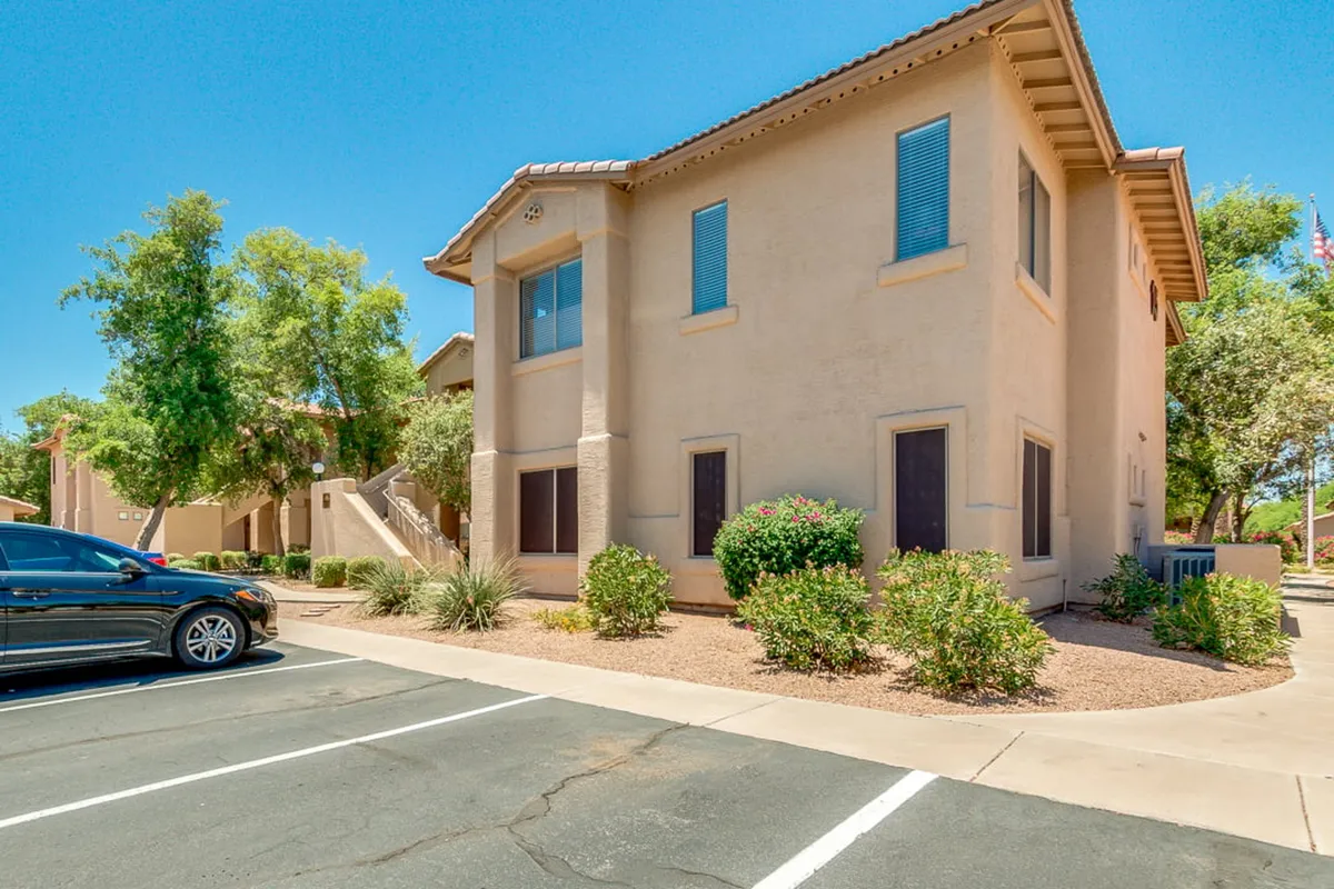 This image displays the two-story building with manicured gardens and a visible parking lot, situating the condo in a friendly, accessible neighborhood. The lush landscaping and community atmosphere are prominent, emphasizing the welcoming environment just steps from your door.