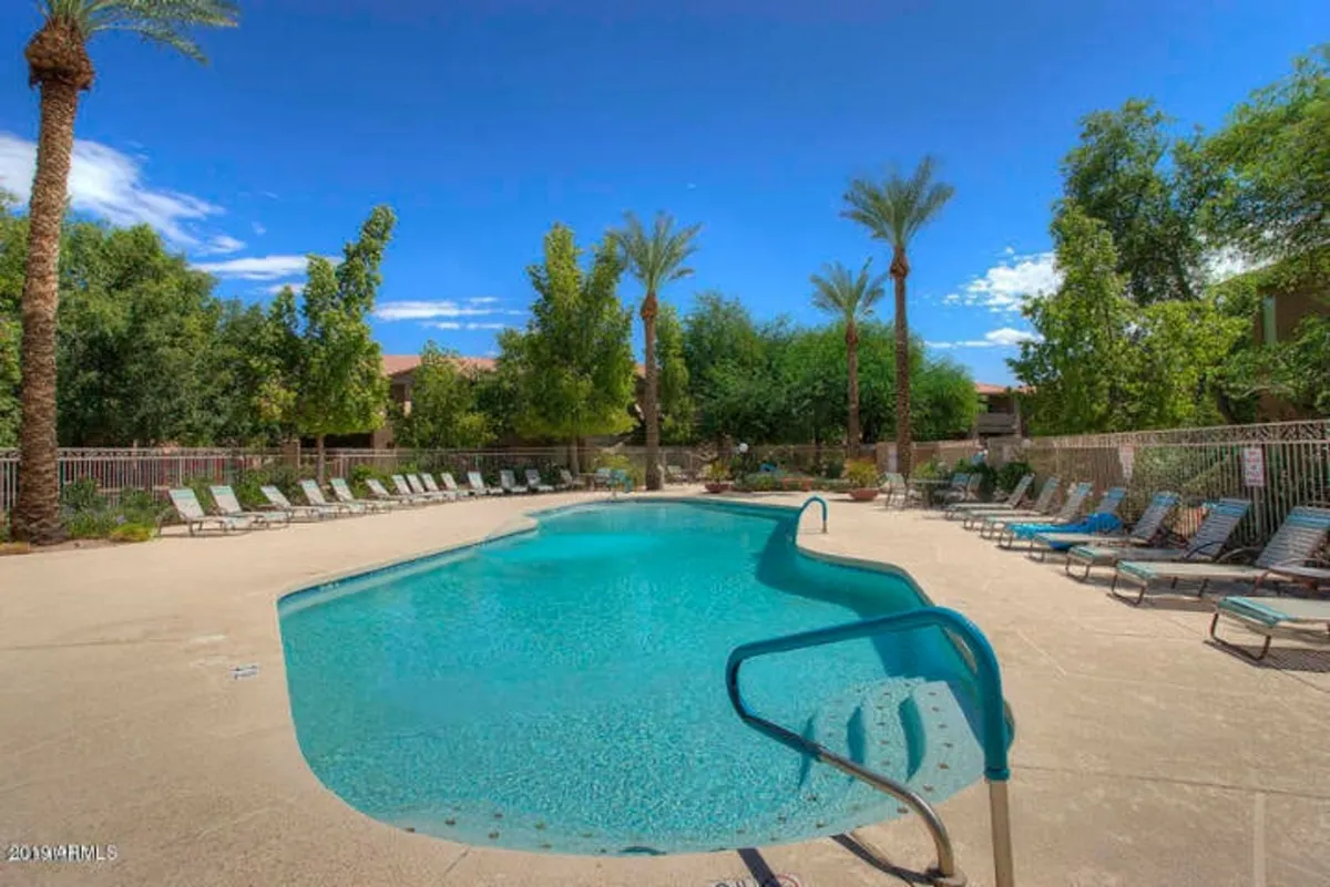 A sparkling heated swimming pool is surrounded by comfortable lounge chairs and lush palm trees. The vibrant blue water glistens under the sun, inviting residents to swim, sunbathe, or simply relax poolside. The atmosphere is relaxed and vacation-like, echoing the carefree mood of a resort.